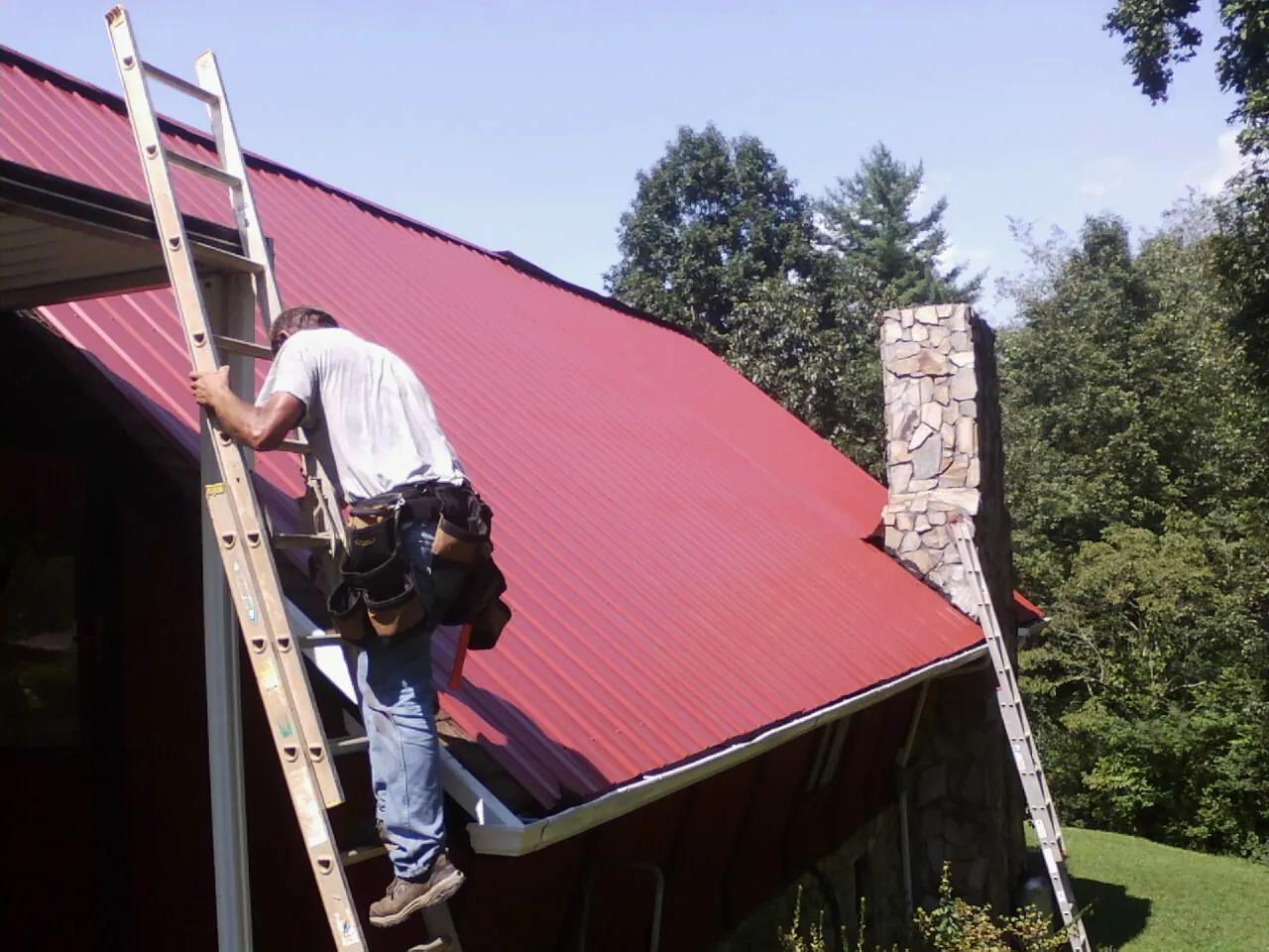 Roofer on a ladder beside a red metal roof and stone chimney — Soffit & Fascia Repair by Roofing Brothers in Mohnton
